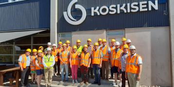 Outdoor group photo from a factory visit, featuring around 30 people posing while wearing orange vests and yellow helmets.
