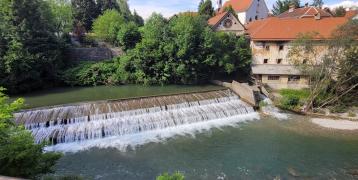 A picturesque view of a small waterfall and river in Škofja Loka, Slovenia, surrounded by lush greenery and historic buildings with red-tiled roofs. The clear blue sky and serene landscape provide a peaceful atmosphere.