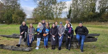 A group of ADAPTO project partners sits together on a large wooden bench outdoors at Cabragh Wetlands, Ireland. Behind them are green trees and an open field. The group, dressed warmly for autumn, is smiling and holding informational brochures.