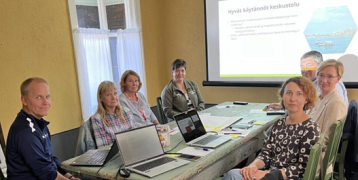 Participants of DETOCS Regional Stakeholder Meeting sit at a table with laptops and a presentation
