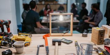 Τhe picture shows a bench with tools and behind it a group of people repairing things