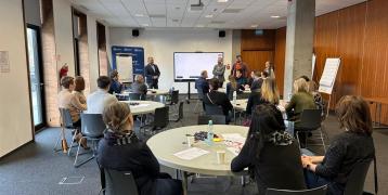 Participants sit in groups during a workshop, with facilitators presenting at a whiteboard in a bright, modern room.