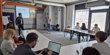 People attend a meeting where two men present the “Płock Connect” migrant app. Participants sit around tables with laptops and notes.