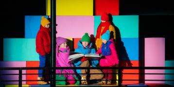 Five children and young people in colorful clothing reading large book on stage