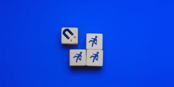 Four wooden blocks are arranged on a bright blue background. One block features a magnet icon with red lines indicating attraction, while the other three blocks each display a businessperson figure walking toward the magnet.