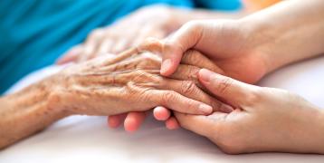 The image depicts a close-up of two hands: one is older, showing visible wrinkles and age spots, and the other is younger, gently holding the older hand. In the blurred background, a person is lying down, covered with a blue blanket or garment. This scene conveys a sense of care and connection between generations.