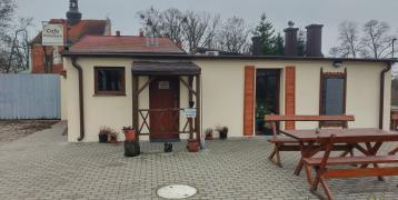 The image depicts a small café called "Cafe Francesco," which has been established in former farm buildings belonging to the parish of the Crucified Jesus and Our Lady of Sorrows in Pakość, located on Kalwaria Hill. What makes this café unique is that visitors can enjoy it free of charge. In return for the hospitality, they have the option to leave a donation of any amount as a token of appreciation.