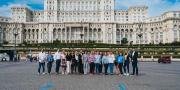 A group of participants from the SHARING project event poses in front of the imposing Palace of the Parliament in Bucharest, Romania, under a bright blue sky. The group consists of men and women standing on a cobblestone square, facing the camera, with some smiling and engaging with each other. In the background, the massive white neoclassical facade of the building stretches across the scene, adding grandeur and context to the gathering.