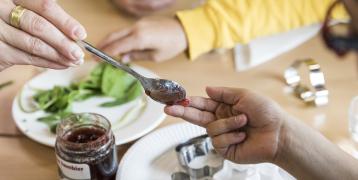 A close-up of two hands during a shared cooking activity—one adult holds a spoon with homemade jam, gently placing a small amount onto a child's fingertip.