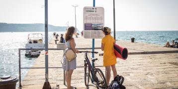 The image shows cyclists waiting for a boat, chacking the timetable. The boat provides transport for passengers and bicycles, complementing the offer for cyclists.