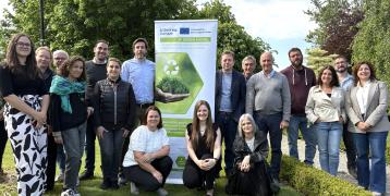  group of 18 people, both men and women of varying ages, stand and kneel on grass outdoors in front of a vertical banner. The banner displays logos of the Interreg Europe program and the European Union, and it promotes the "3F GREEN MODEL" project focused on circular economy and sustainable forest value chains. The group is smiling and appears to be part of a professional or collaborative environmental initiative. The background features lush green trees and a partly cloudy sky.