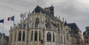 A Gothic-style cathedral with pointed arches, ornate spires, and large stained glass windows, seen from a plaza with parked cars. Three flags—EU, French, and a local flag—stand in the foreground, suggesting a European town setting