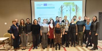 A group of individuals standing in a conference room in front of a projector screen displaying a slide for the "2nd International Partnership Meeting" of the FBI4EU project. The background slide shows the Interreg Europe logo and an illustration of a forest.