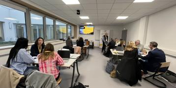 A group of people are engaged in discussions around tables in a bright, modern classroom. Some are working on laptops while a presenter addresses the group at the front.