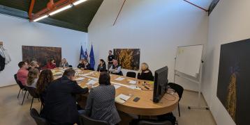 People are seated around the table while the facilitator stands and guides the discussion.