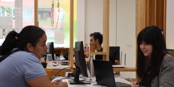  The image shows an individual consultation at the Valencia Energy Office. On the left, a woman is sitting at a desk speaking with another woman on the right, who is smiling while holding a pen and reviewing a document. Both are seated in front of computers in a modern, open-space office setting with light wooden elements and plants hanging from above, creating a welcoming environment. The consultation is focused on energy-related advice.