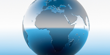 A large inscription ‘18 December, International Migrants Day’ is visible at the top, with a graphic of a globe below, under which a number of silhouettes of migrants - adults and children carrying luggage - are visible.