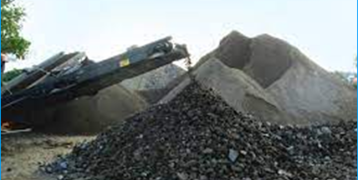 A mechanical crusher discharges a pile of mixed aggregates, featuring both larger dark, rocky materials in the foreground and finer sandy aggregates in the background. The machine's conveyor is visible to the left, depositing crushed materials, with large heaps of sand and gravel behind it. This scene depicts the process of crushing and sorting raw materials into different grades for use in construction. Trees and natural surroundings frame the industrial activity.