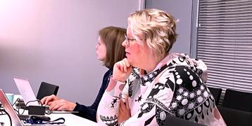 Light-haired woman sitting beside the meeting room table looking down to her laptop and giving presentation.