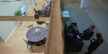 The image shows the test apartment viewed from above, with two people sitting inside the apartment and having a conversation. Outside the apartment, two people are observing the activity inside on monitors.