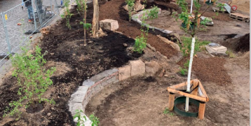 The fotograph shows a square that has been unsealed. Where there used to be asphalt, there are now beds for flowers, shrubs and vegetables as well as newly planted trees. 