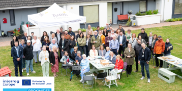 Group photo of PROXIMITIES study visit participants and local residents gathered outdoors at Arus Mhuire, an age-friendly housing scheme in Cork. People are smiling and standing or sitting around picnic tables, under a Tuath Housing canopy. PROXIMITIES and Interreg Europe logos are visible in the foreground.