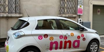 NINA Neighbourhood car sharing NINA car sharing vehicle, a white car with a pink and red NINA logo on the side and the words ‘neighbourhood car sharing’, parked in a dedicated car park indicated by a sign.