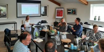 A group of stakeholders engaged in a discussion during the GIFT project Regional Stakeholder Group meeting in Fryslân, July 2024. A presentation is displayed on a screen, and participants are seated around a conference table with laptops, coffee, and refr