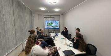 PROXIMITIES stakeholders from the Riga Planning Region discuss a presentation on "PROXIMITIES Olaine" in a meeting room. The screen displays project visuals, and the table holds coffee cups, water glasses, and notebooks.