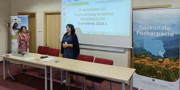 A woman standing behind a table speaks in a closed room. Behind a powerpoint screen and on the side another woman standing and listening.