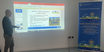 Eddie Meegan presenting climate adaptation strategies during the SustainableTipp meeting, showing a slide on the ADAPTO project, with a climate action banner beside him.