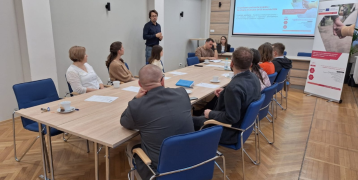People can be seen sitting at tables set together watching a presentation projected on a screen at the back of the room. On the left, between the tables and the wall, stands a person in the middle of a speech.  To the right, a rollup with a poster of the SIRM project is visible.
