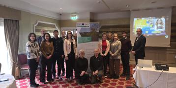 In the picture, a diverse group of men and women from the INSPIRE project stands together, posing for a photo. They are gathered after a Regional Stakeholder meeting at a facility in Spruce & Willow, located in Ardpatrick, Co. Limerick. The group smiles and stands proudly in front of a large banner displaying the INSPIRE project logo, highlighting their collaboration and the purpose of the event. The setting appears professional, with the modern facility in the background, offering a sense of teamwork.