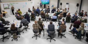 A forestry conference in Norddjurs, Denmark, with participants seated at round tables listening to a speaker presenting on a large screen. A facilitator is writing on a whiteboard in the background. The event takes place in a modern indoor setting with professional equipment.