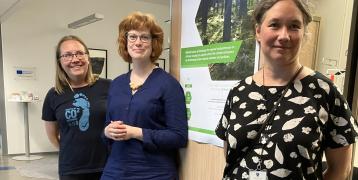 Three smiling women are standing and leaning against the meeting room wall.