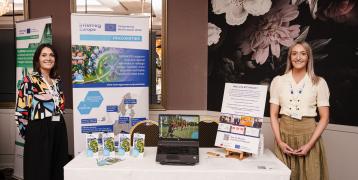 Two women stand on either side of a promotional display table for the Interreg Europe PROXIMITIES project at an indoor event. Behind them are banners showcasing the PROXIMITIES project and the ETC Network. On the table are brochures, a laptop playing a video, and informational materials. The setting appears to be a conference or exhibition space with floral wallpaper and elegant decor.