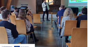 A speaker stands at the front of a room pointing toward a presentation slide on a screen during a Riga Planning Region Working Group meeting. Attendees are seated in rows facing forward. The setting is a restored wooden building with high ceilings and natural light. The PROXIMITIES and Interreg Europe logos appear in the corner.