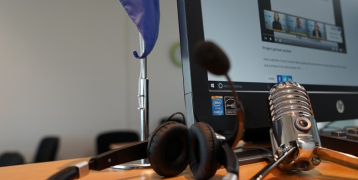 Professional tools for creating a video Close-up of a desk setup for virtual meetings or online collaboration. The desk features a European Union flag, a computer monitor displaying a video call or webinar interface, a pair of black headphones, and a vintage-style microphone.