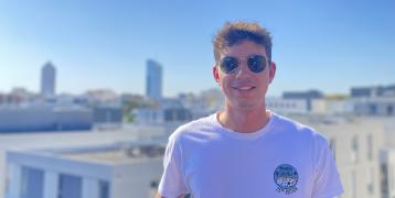 Dark-haired student boy with sun glasses and white T-shirt smiles at the rooftops of Lyon.