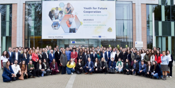 A large group of young people poses for a group photo in front of a modern glass-fronted building. Above them, a large banner reads "Youth for Future Cooperation – on the road to Interreg GO!", with event details indicating gatherings in Brussels (November 15, 2024) and Gorizia-Nova Gorica (March 27-28, 2025). The banner also features images of young professionals engaging in discussions and collaborative activities. The setting appears to be an official EU institution, with the European Commission logo vis