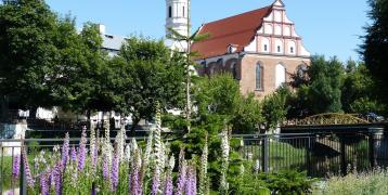 The image shows a church with a tall spire and brick walls, set among trees and colorful flowers in the foreground. A small bridge can be seen in the background, enhancing the peaceful scene