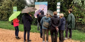 A group of people holding umbrellas stand outdoors in front of an information board and signage, examining a map and illustrations on biodiversity. The group is dressed in rain jackets, indicating rainy weather, with green foliage and a gravel path in the background. The scene appears to be part of an outdoor learning session or study visit focused on environmental conservation and green infrastructure.