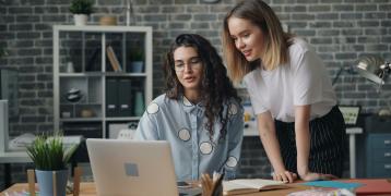 Two women collaborate at a desk in front of a computer